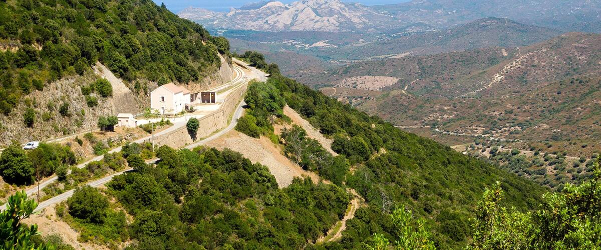 A winding road through a mountainous landscape with a small building on the left and a view of distant mountains and valleys.