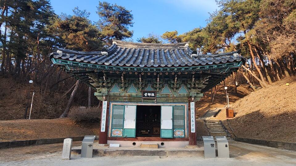 A traditional Korean pavilion with a tiled roof, surrounded by trees and a dirt path.