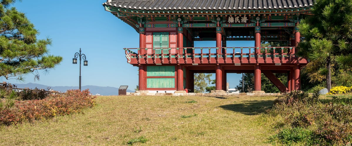 A traditional Korean pavilion with a tiled roof, surrounded by grass and trees.