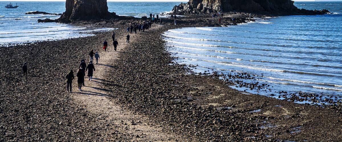 A rocky beach with people walking, two prominent rock formations, and calm sea.