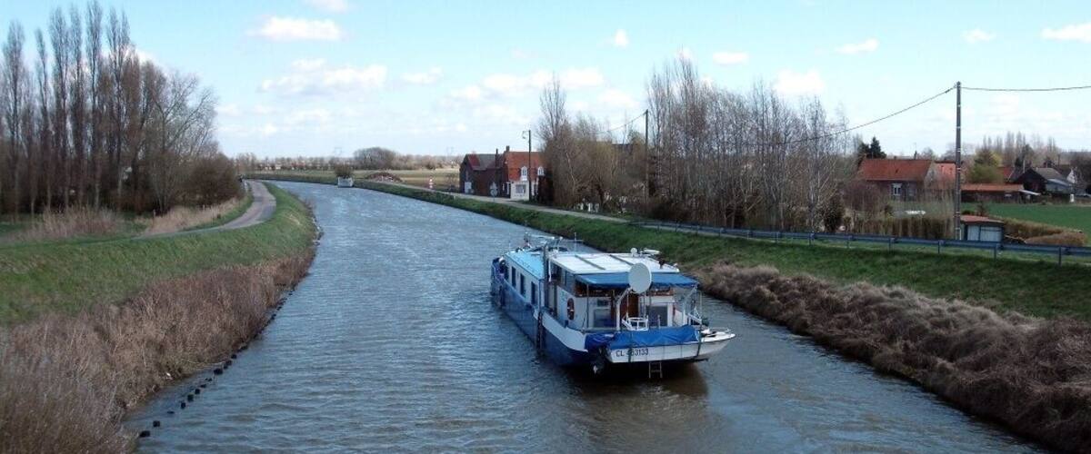 A narrow waterway with a blue boat, surrounded by grassy banks and leafless trees.