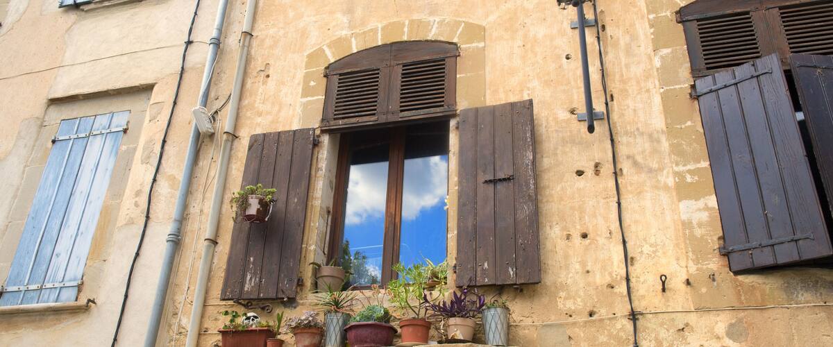 A building with a window with wooden shutters, a street lamp, and multiple potted plants on the windowsill.