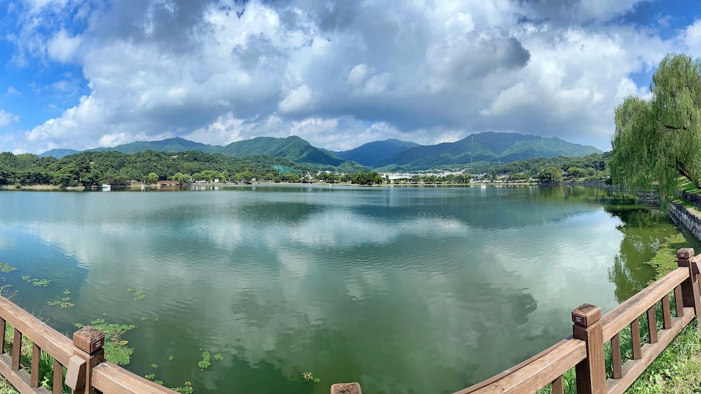 A calm lake with a wooden fence, green trees, and mountains in the background.