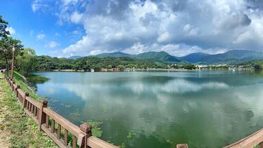Um lago calmo com uma cerca de madeira, árvores verdes e montanhas ao fundo.