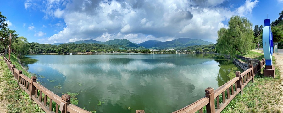 Un lago tranquilo con una valla de madera, árboles verdes y montañas al fondo.