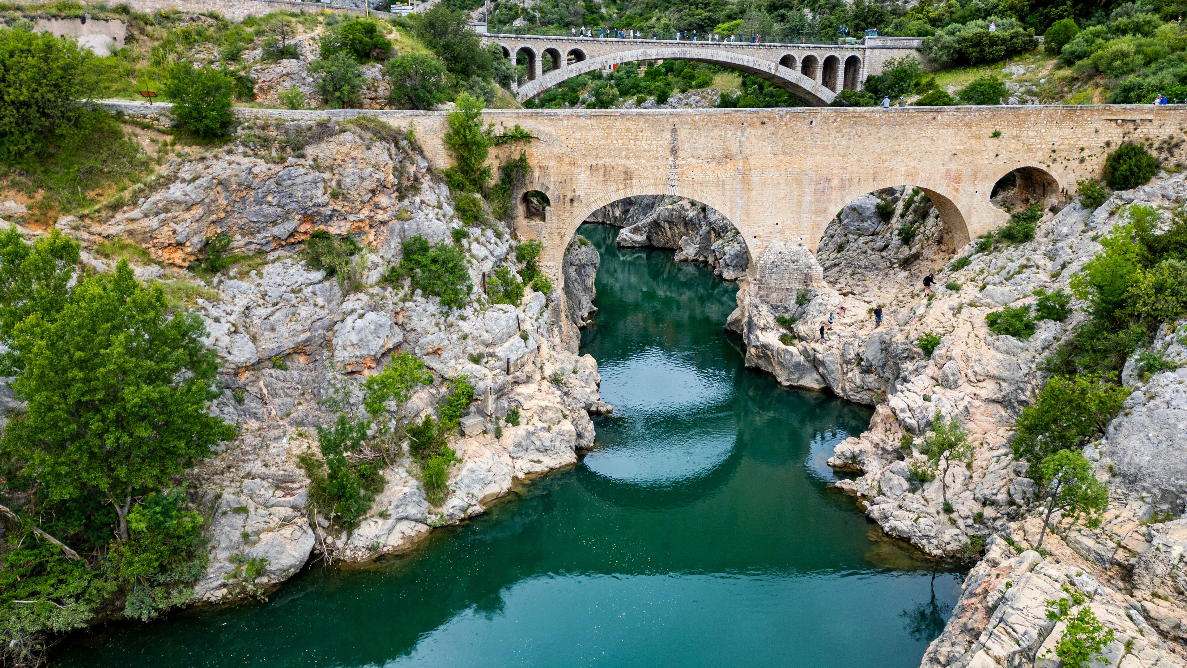 A stone bridge with arches over a river surrounded by rocky terrain and greenery.