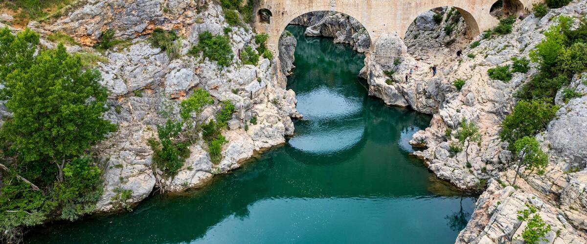 A stone bridge with arches over a river surrounded by rocky terrain and greenery.