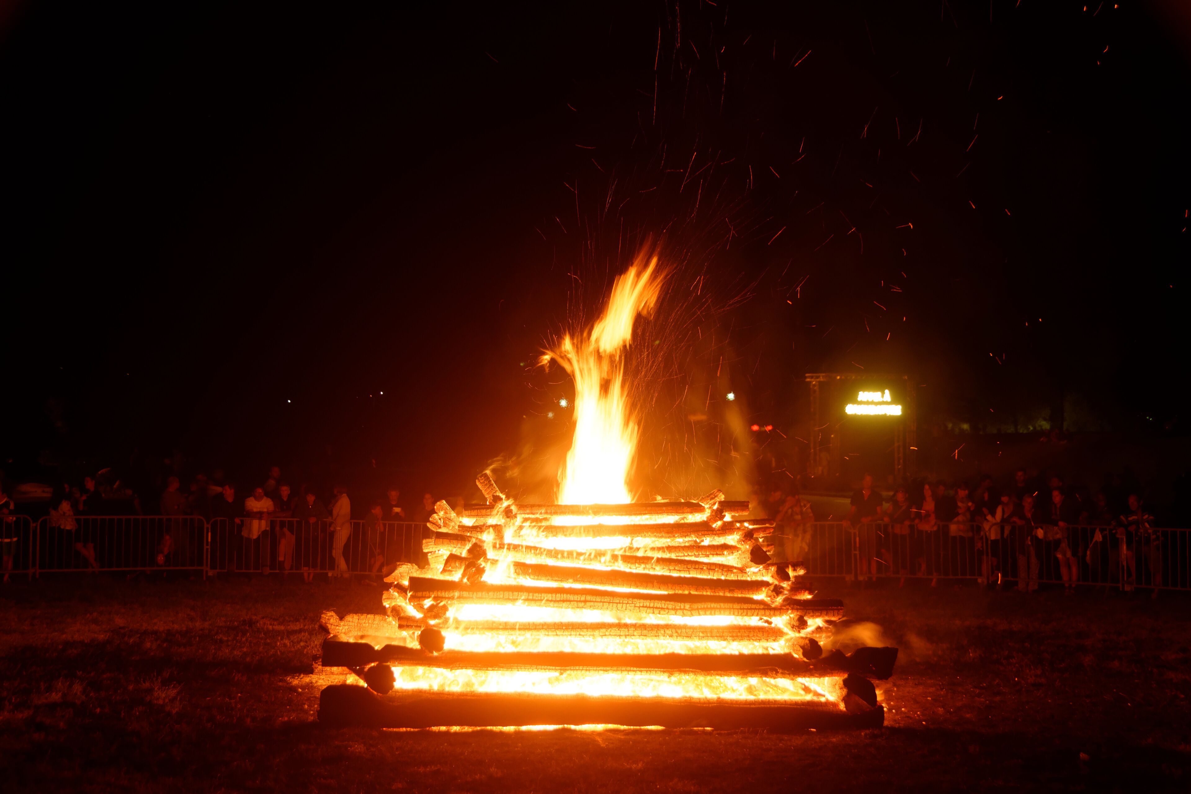 A large bonfire with flames shooting up, surrounded by a crowd of people and metal barriers.