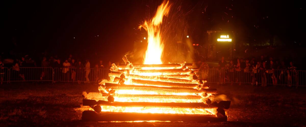 A large bonfire with flames shooting up, surrounded by a crowd of people and metal barriers.