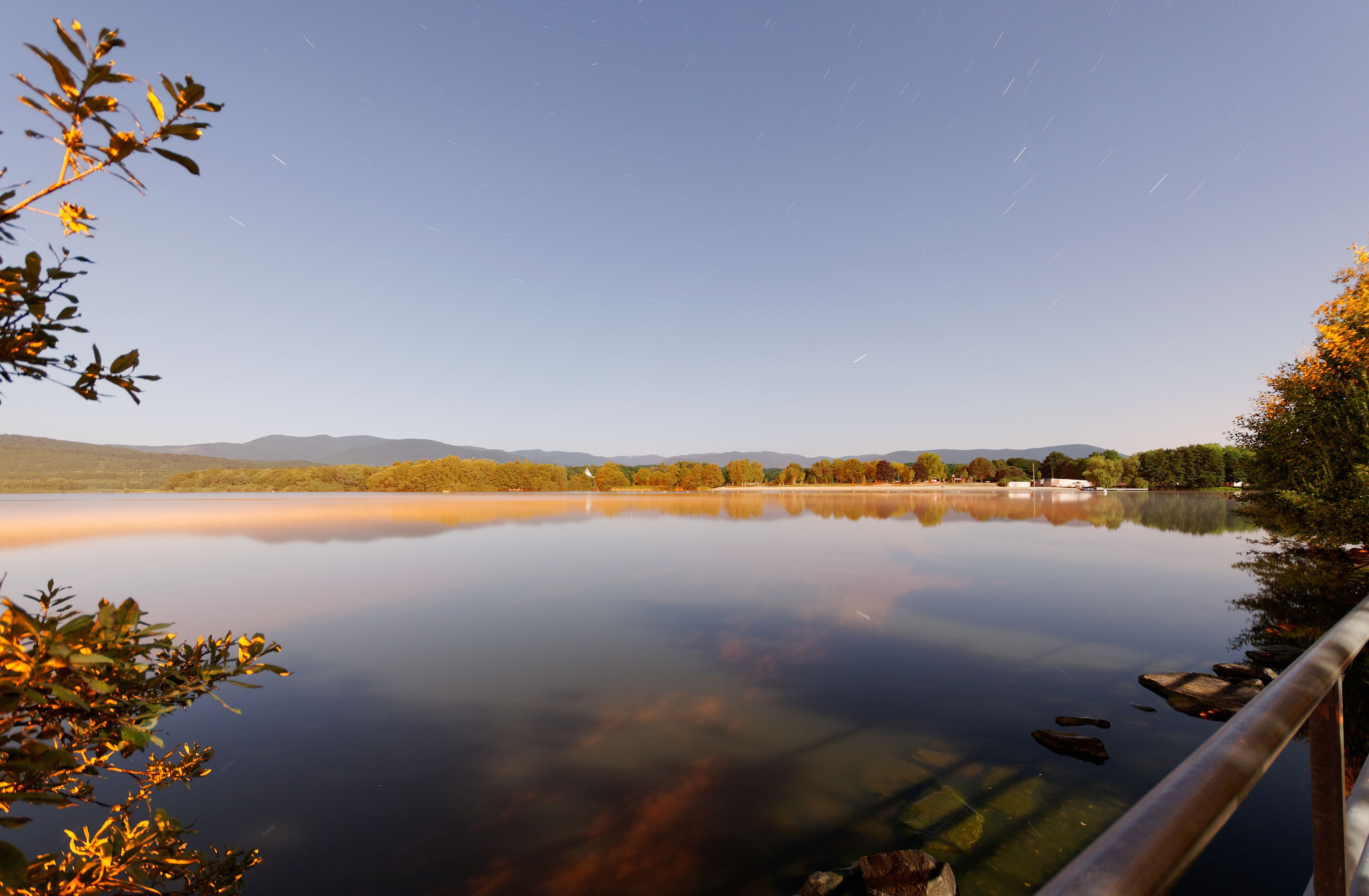 A calm lake with reflections of trees and mountains in the background.