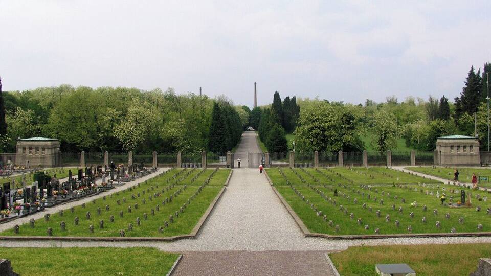 A cemetery with rows of graves, a central pathway, and a large building in the background.