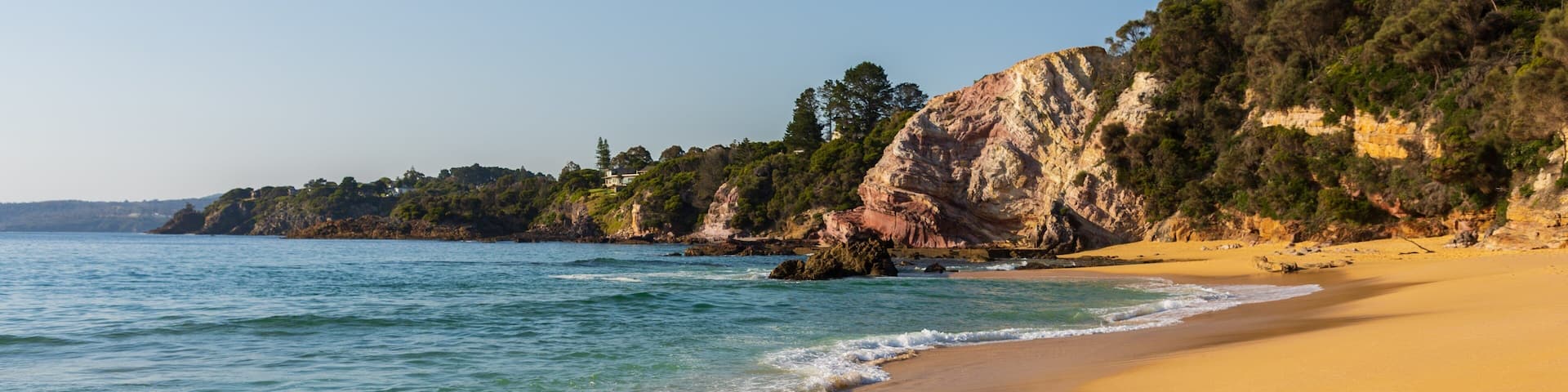A sandy beach with gentle waves, a rocky cliff, and dense greenery in the background.