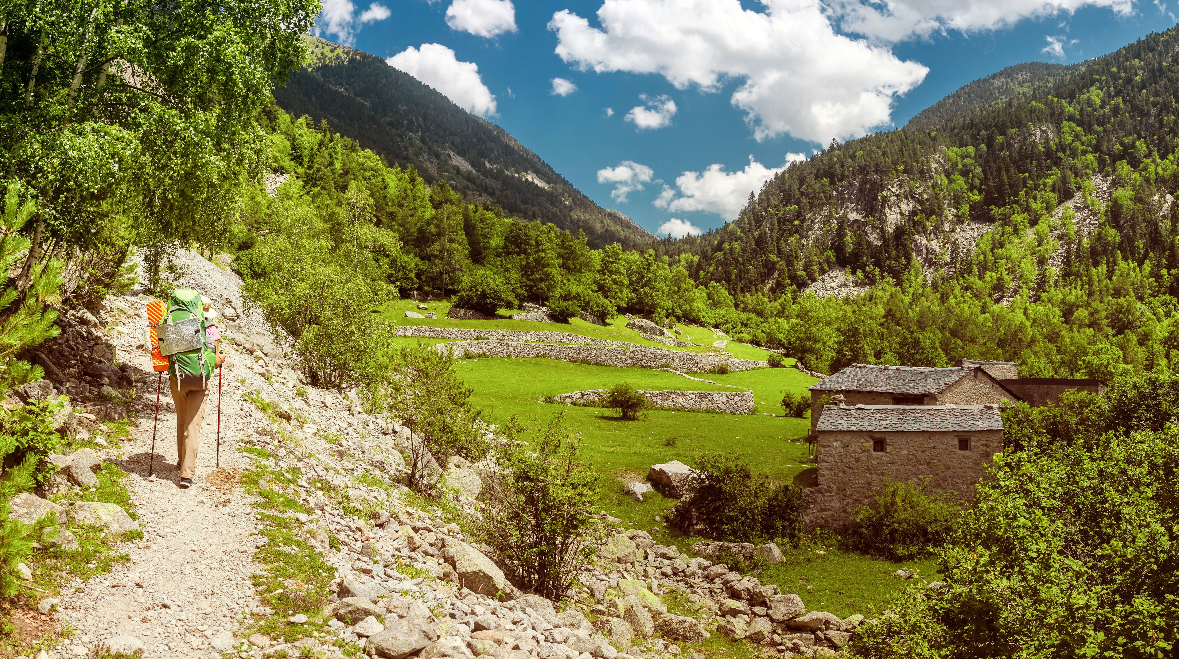 A hiker on a rocky trail, a stone house, and a valley with green fields and mountains.