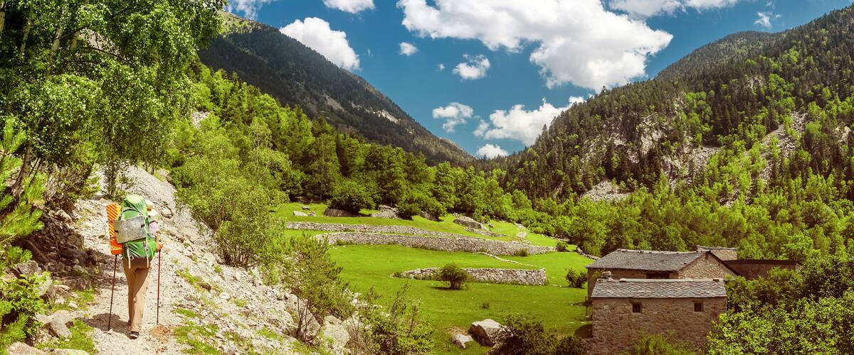 A hiker on a rocky trail, a stone house, and a valley with green fields and mountains.