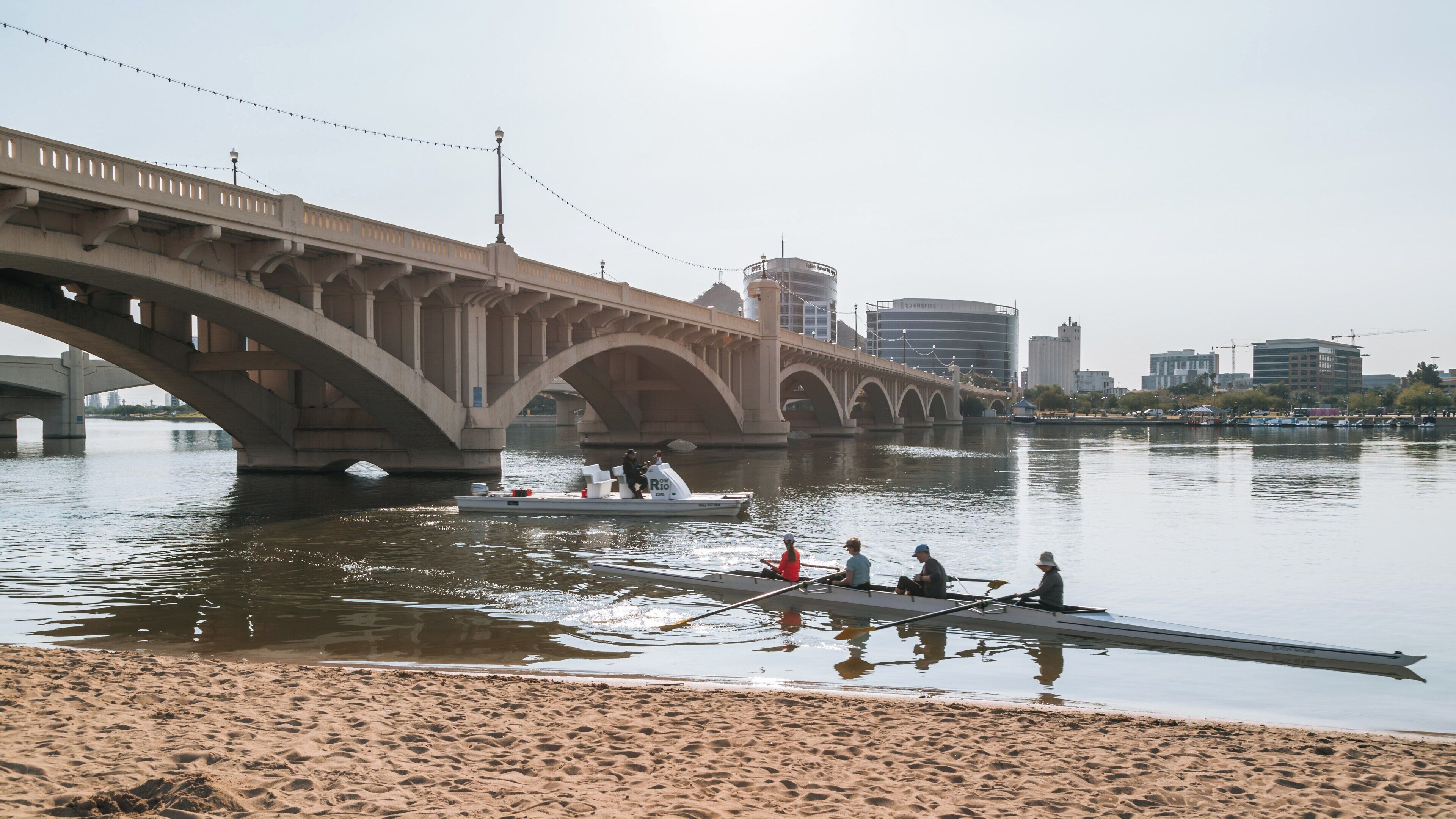 Kayakers navigate Tempe Town Lake under the bridge while urban life thrives in the background on a sunny day in Arizona