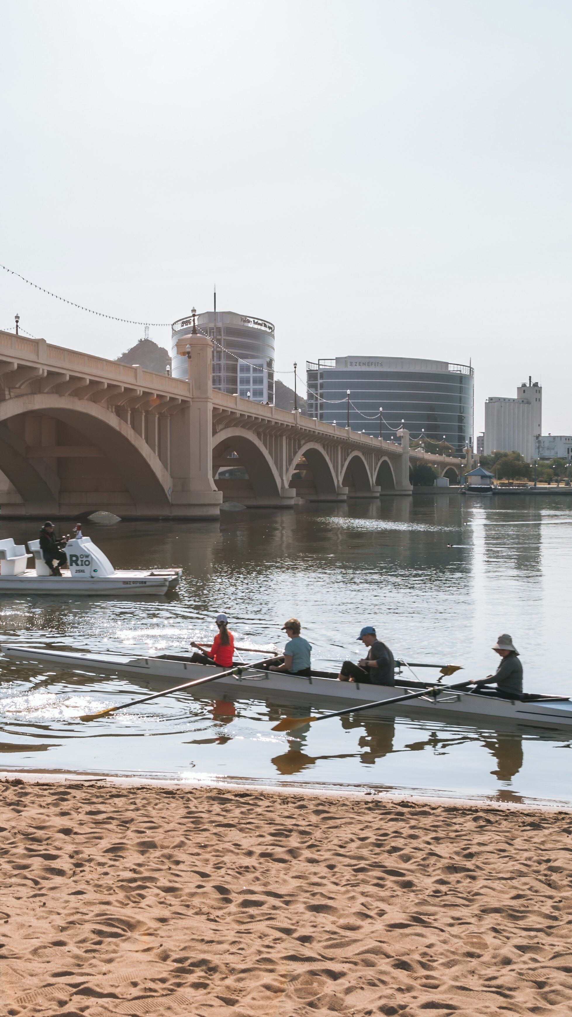 Rowers glide across Tempe Town Lake under a clear sky near urban Tempe, Arizona, showcasing a blend of nature and city life