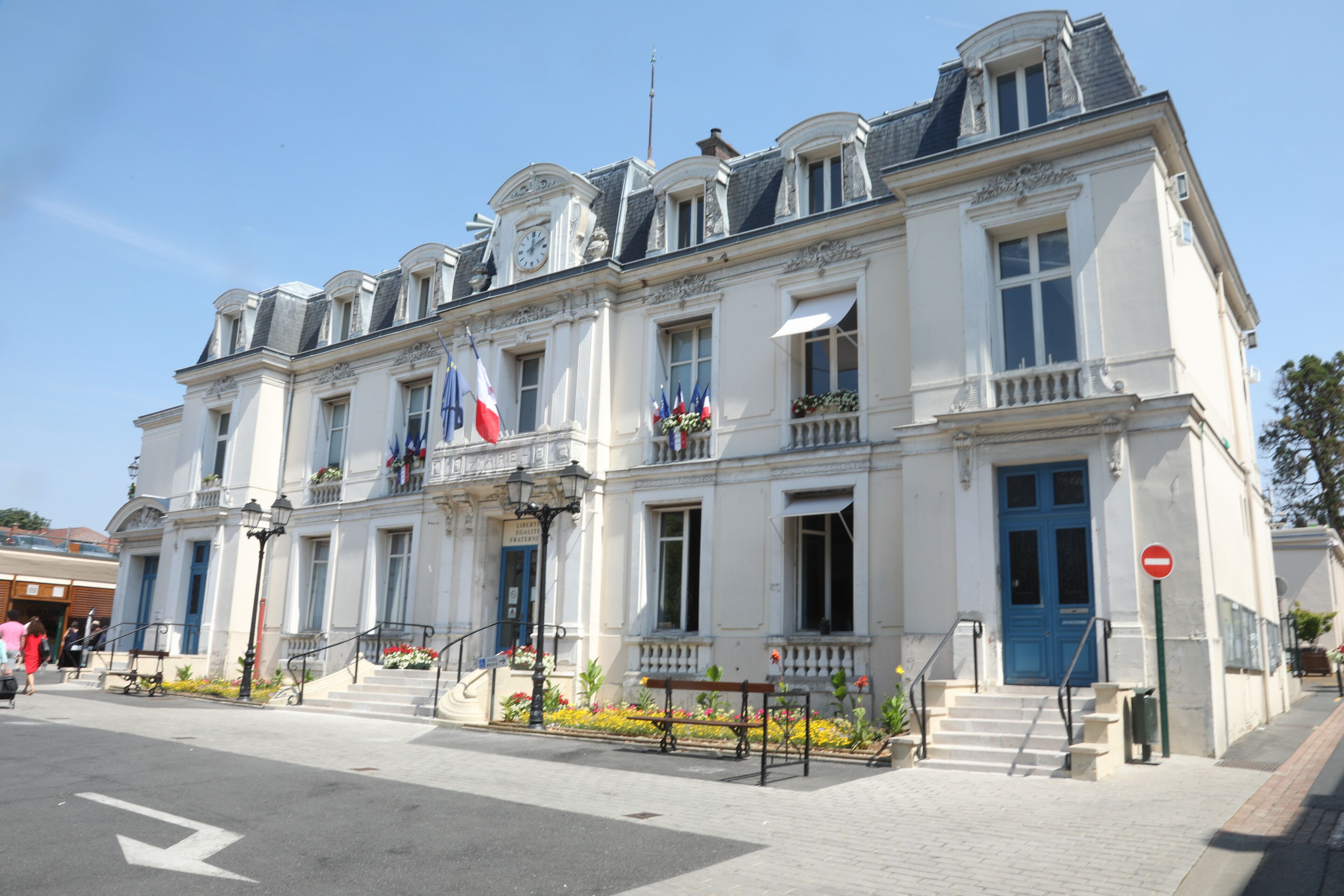 A historic white building with a blue door and multiple flags, including the French flag, on a sunny day.