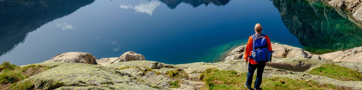 A hiker with a blue backpack on a rocky shore overlooking a clear lake with mountains in the background.