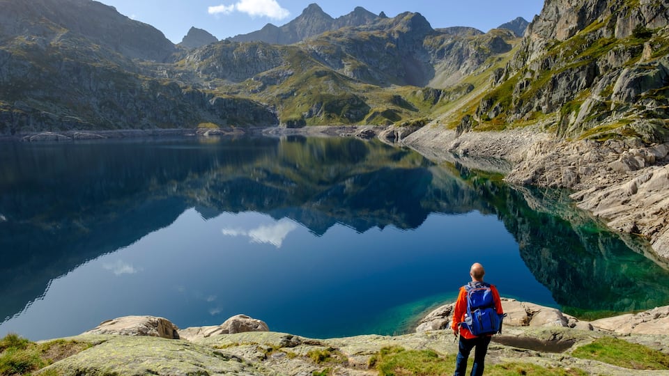 A hiker with a blue backpack on a rocky shore overlooking a clear lake with mountains in the background.