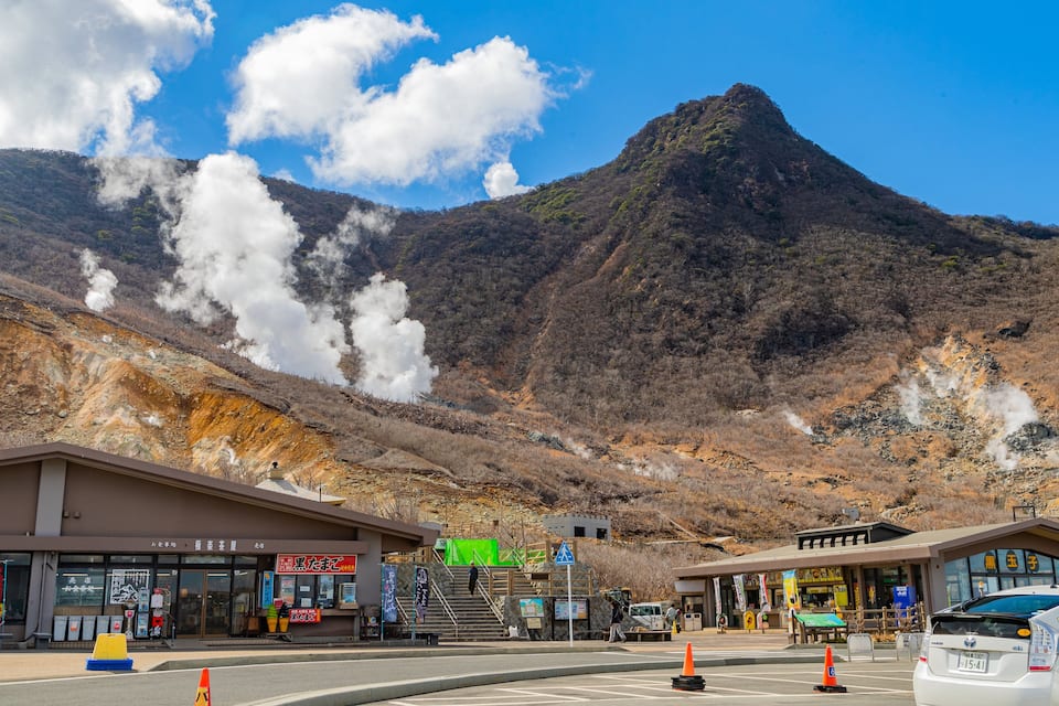 Paisagem montanhosa com um edifício na base, saídas de vapor e um carro branco.