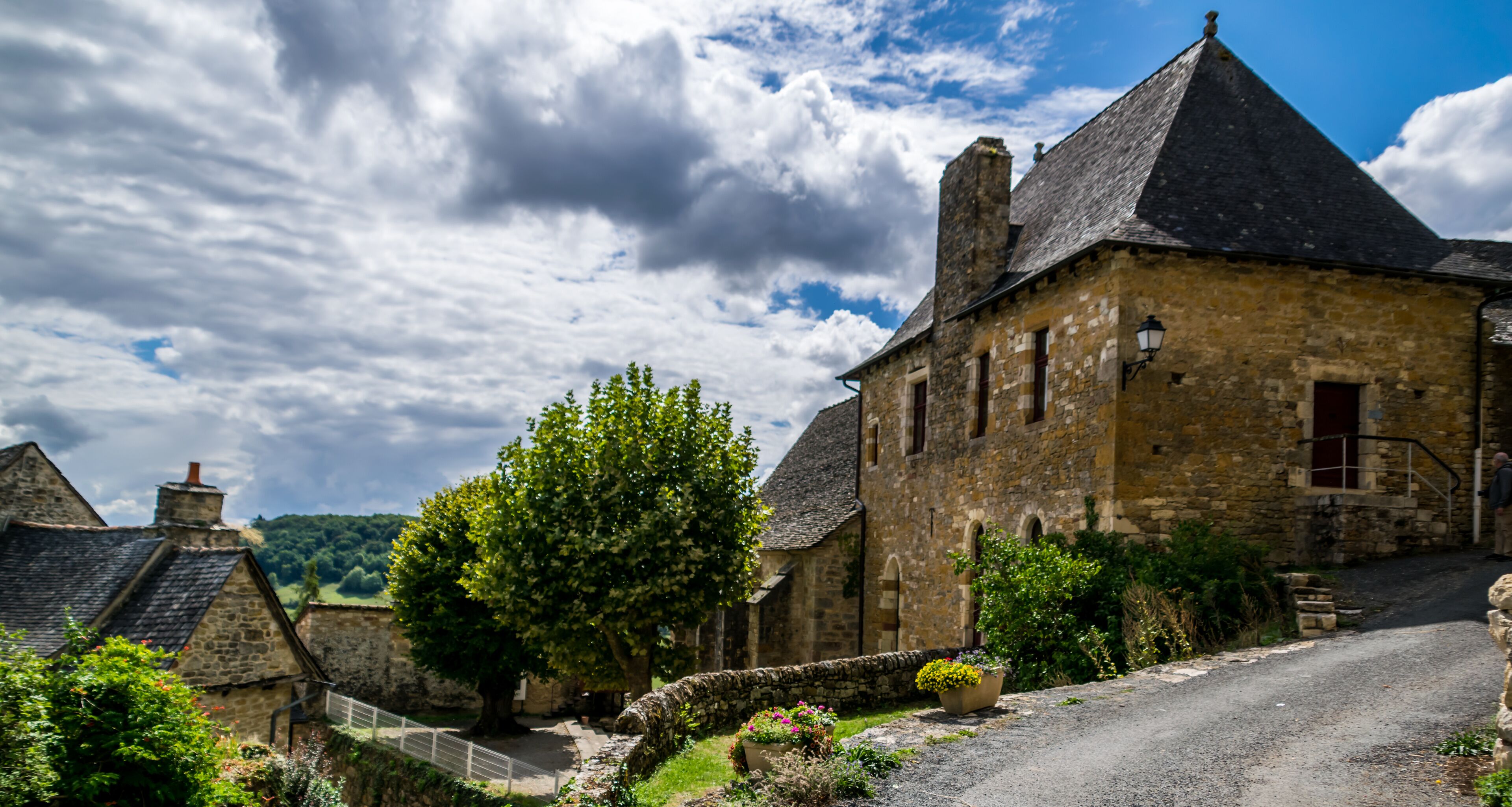 A stone building with a steep roof, surrounded by greenery and a gravel path.