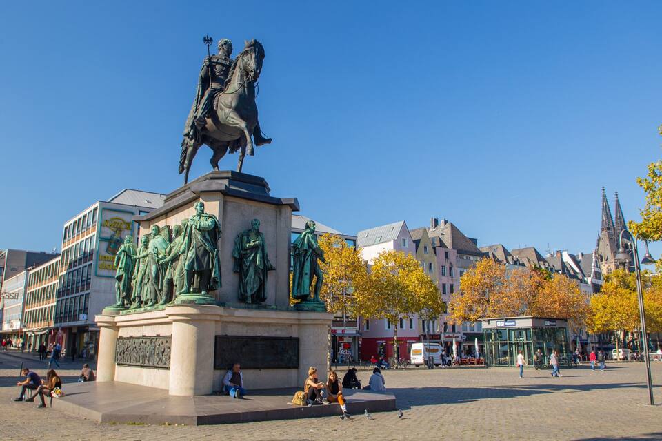 Une statue représentant un homme à cheval, placée sur une place publique, avec des bâtiments et des arbres en arrière-plan.