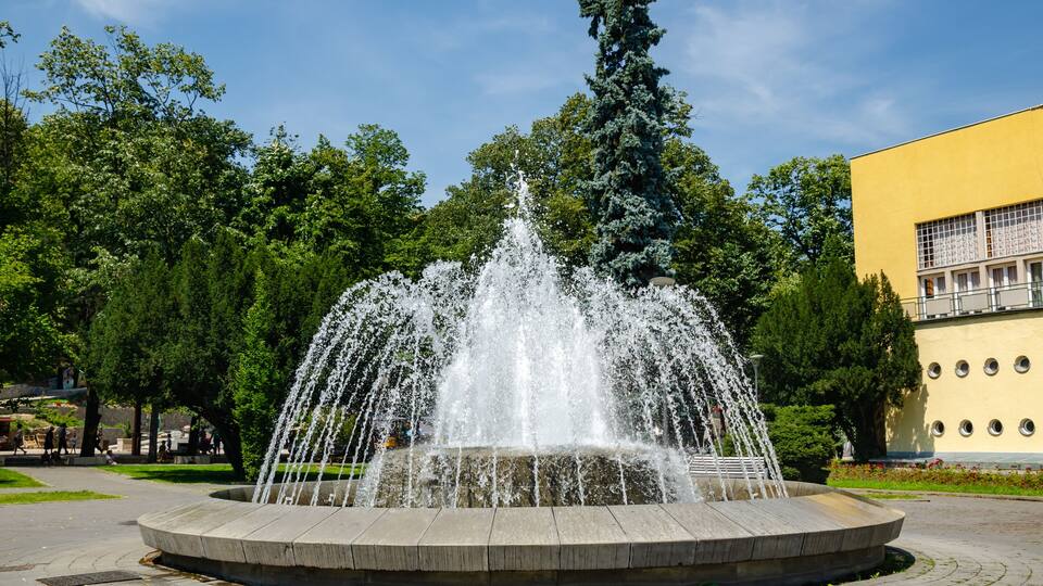 A fountain with water jets in a circular basin, surrounded by trees and a yellow building.