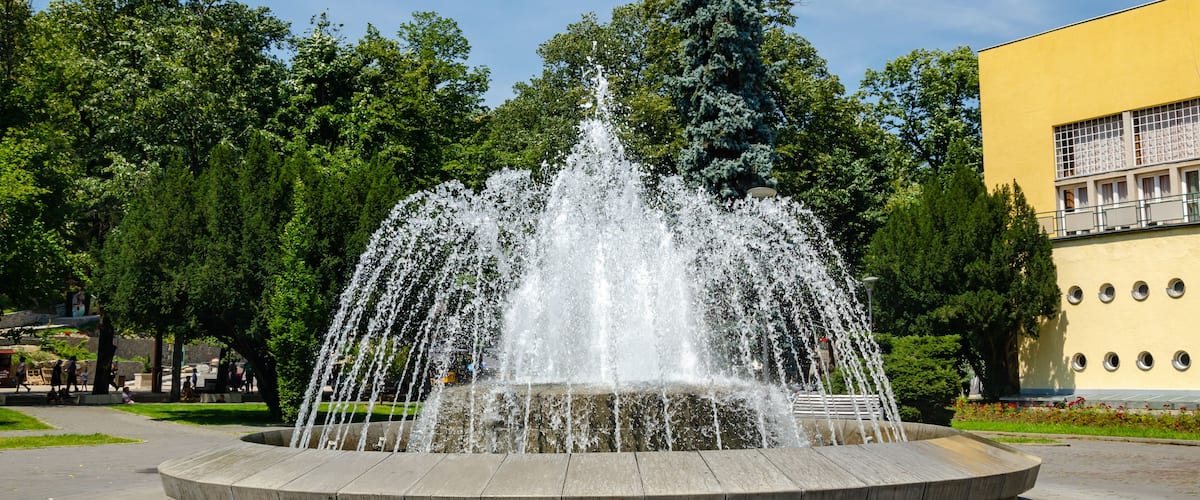 A fountain with water jets in a circular basin, surrounded by trees and a yellow building.