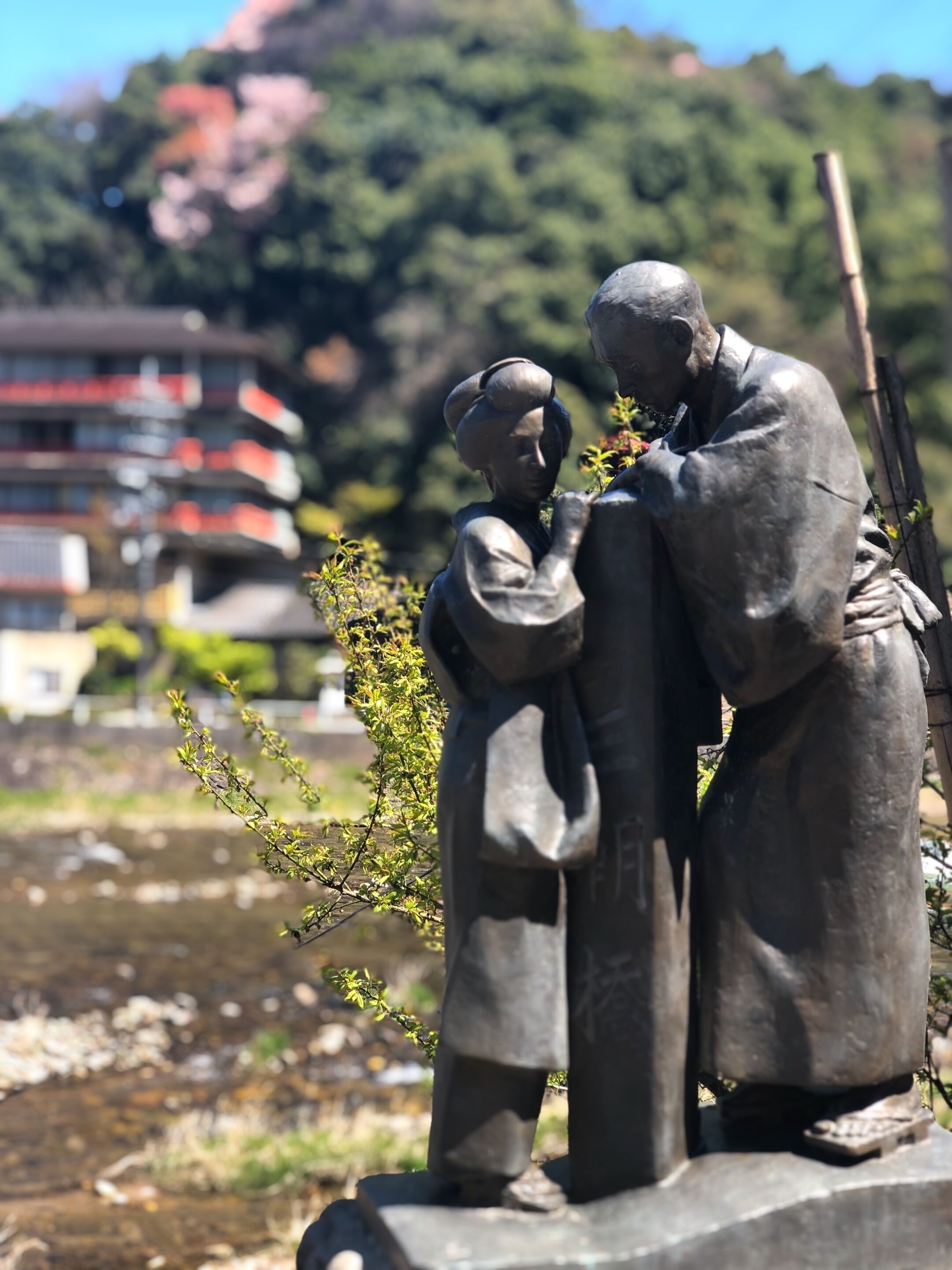 A statue of two figures, one adult and one child, in a park with a building and trees in the background.