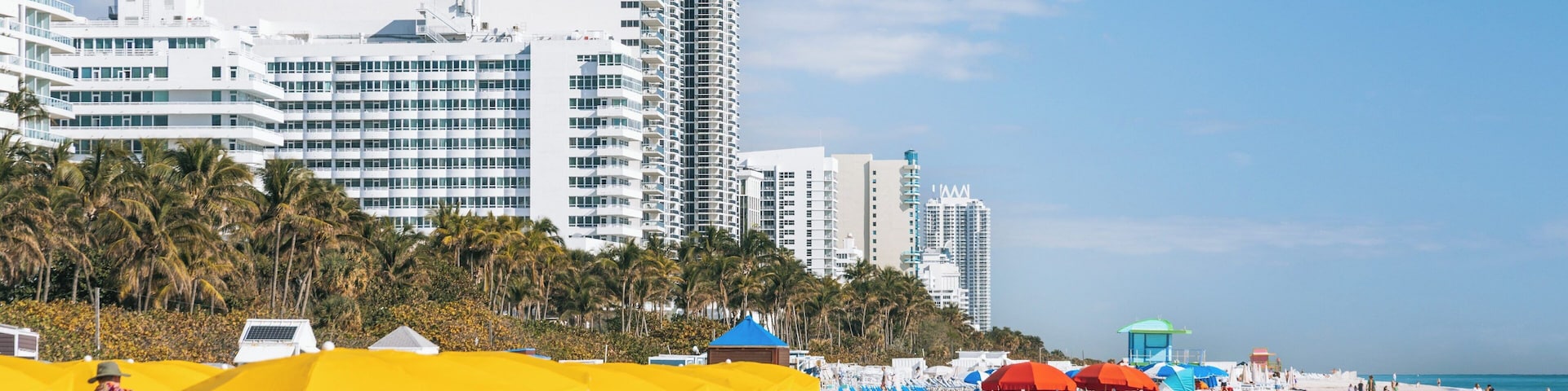 A beach with sun loungers under yellow and red umbrellas, palm trees, and high-rise buildings in the background.