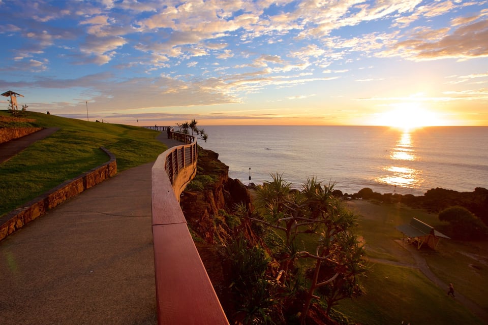 A coastal walkway with a sunset view, grassy areas, and a rocky shoreline.