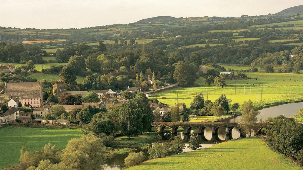 Un paysage rural avec un pont de pierre historique, une végétation luxuriante et une petite ville aux bâtiments d’architecture traditionnelle.