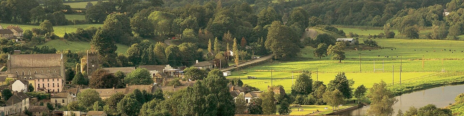 Paisaje rural con un puente de piedra histórico, abundante vegetación y un pueblo pequeño con arquitectura tradicional.