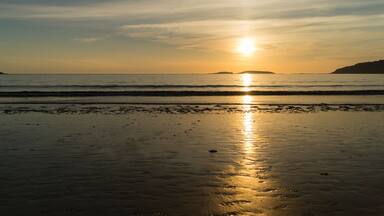 A beach at sunset with the sun reflecting on the wet sand and calm waves approaching the shore.
