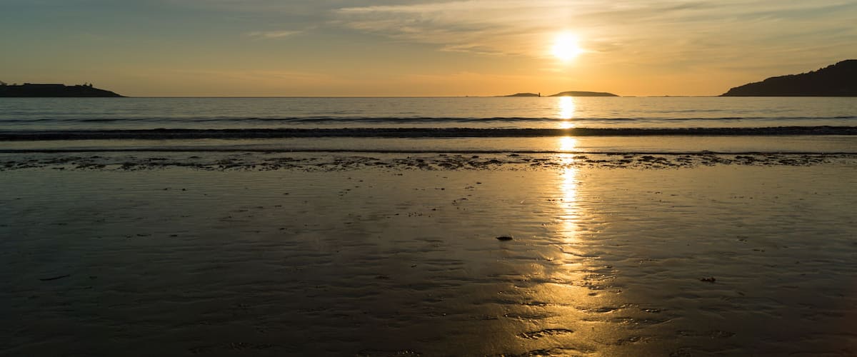 A beach at sunset with the sun reflecting on the wet sand and calm waves approaching the shore.