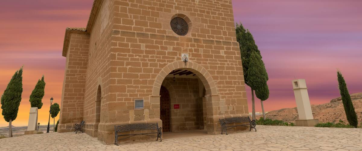 A historic church with a bell tower and arched entrance, surrounded by trees and a cobblestone plaza.