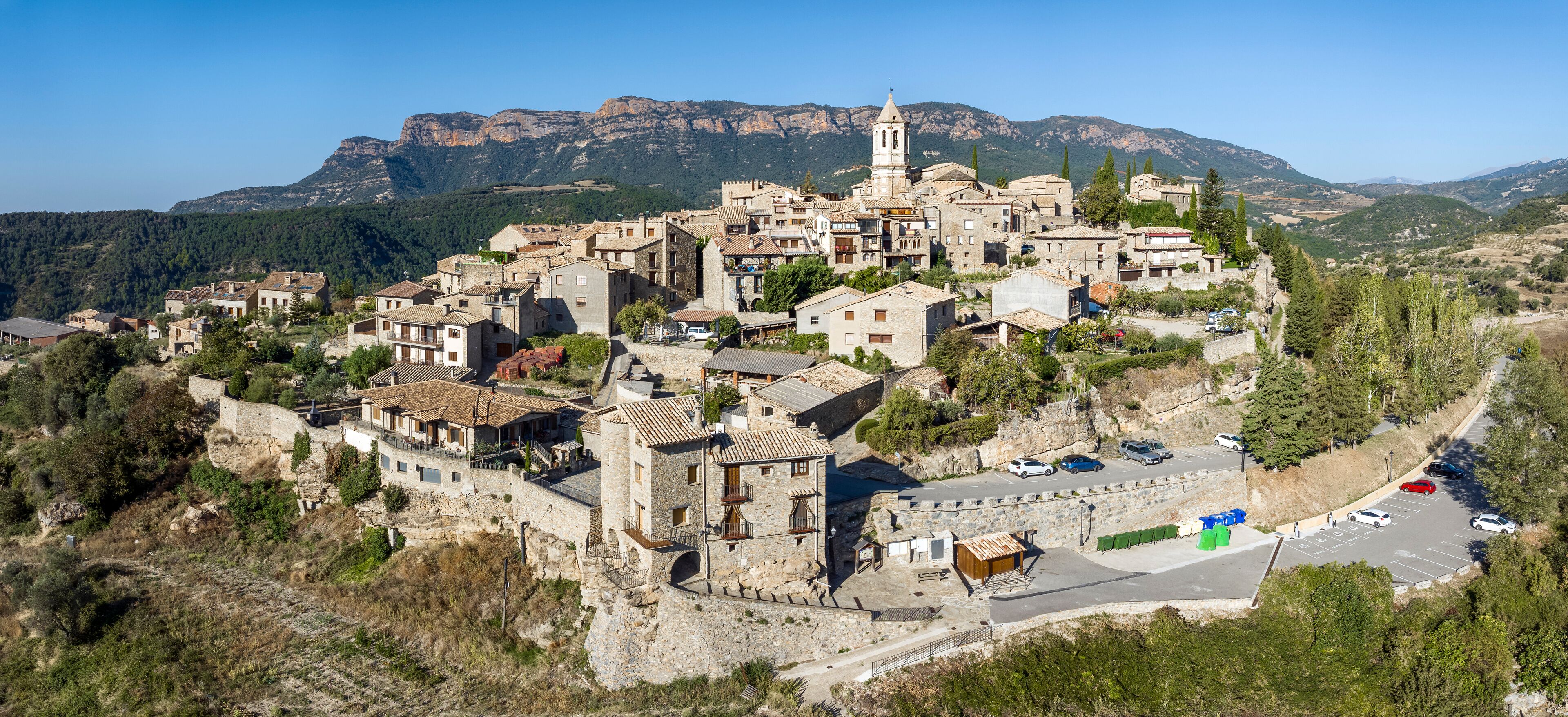A mountainous village with stone buildings, a prominent church tower, and a parking area.