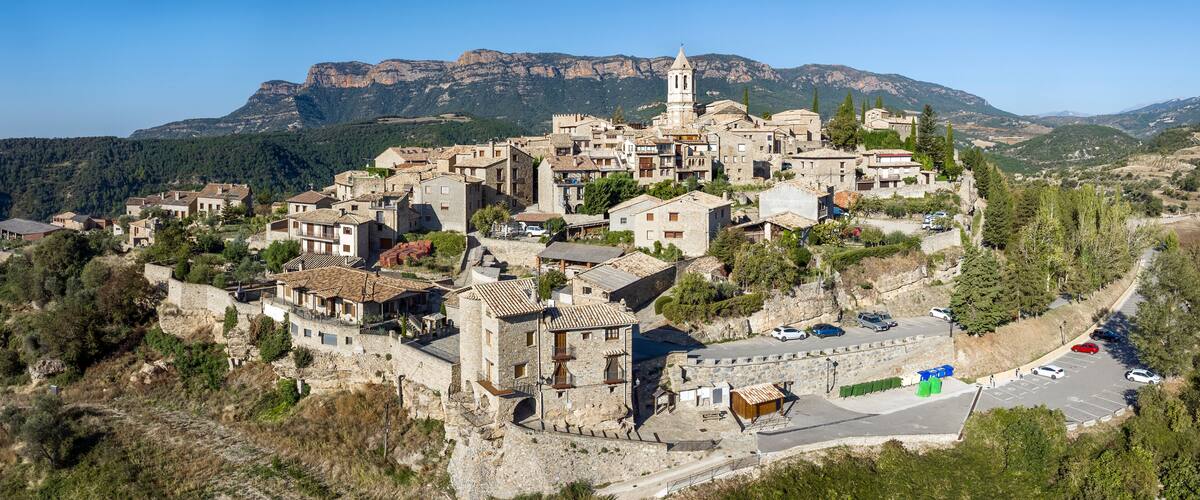 A mountainous village with stone buildings, a prominent church tower, and a parking area.
