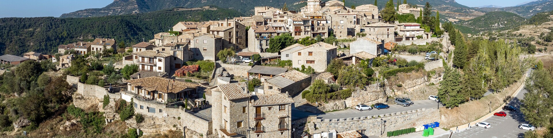 A mountainous village with stone buildings, a prominent church tower, and a parking area.