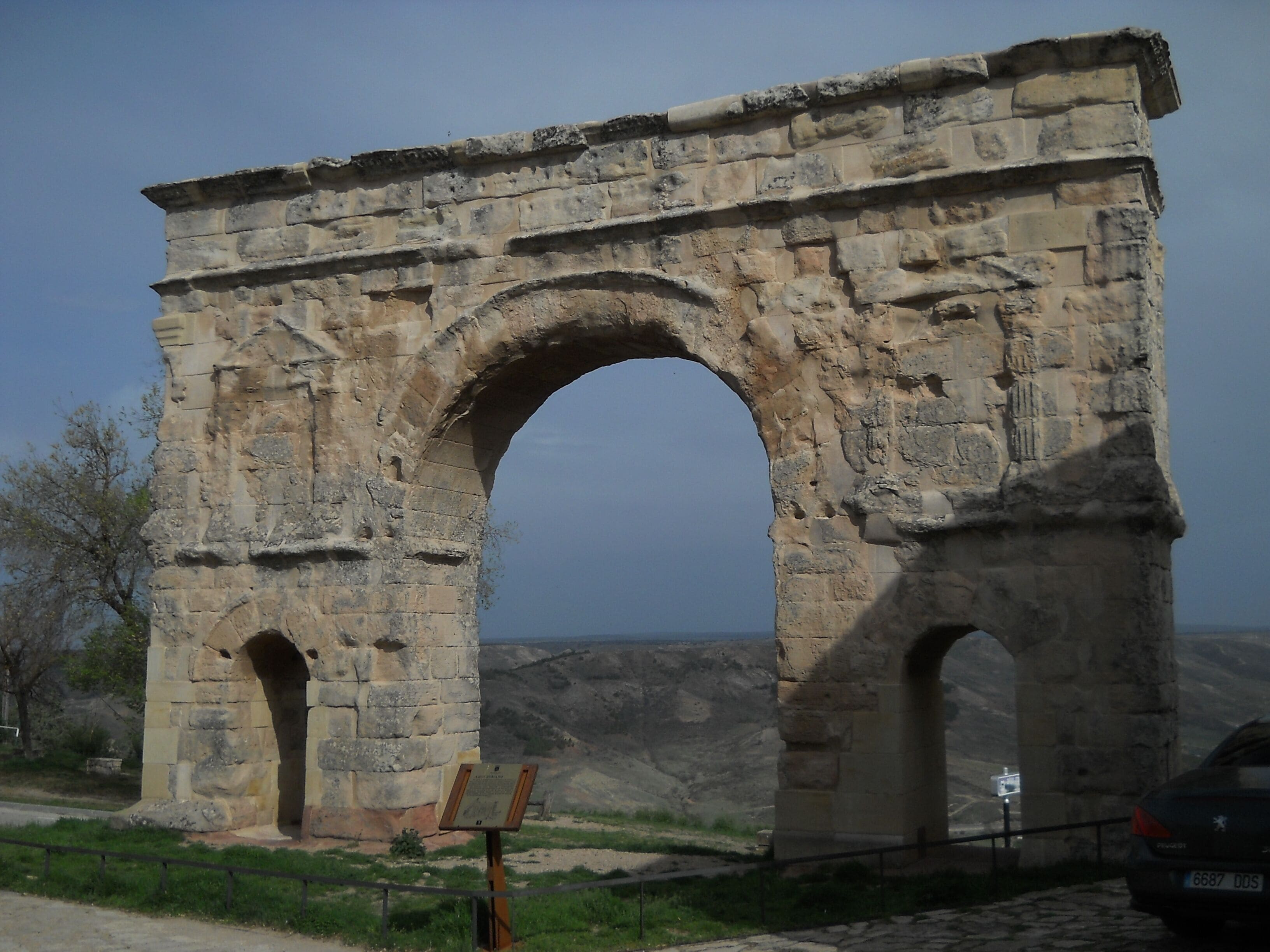An ancient stone archway with two smaller arches on either side, set against a mountainous backdrop.