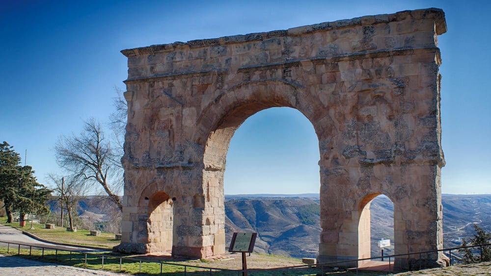 An ancient stone archway with inscriptions, surrounded by a paved area and a mountainous landscape.
