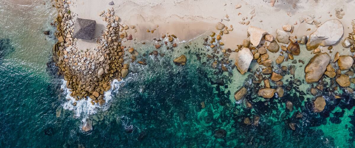 An aerial view of a beach with clear turquoise waters, scattered rocks, and palm trees.