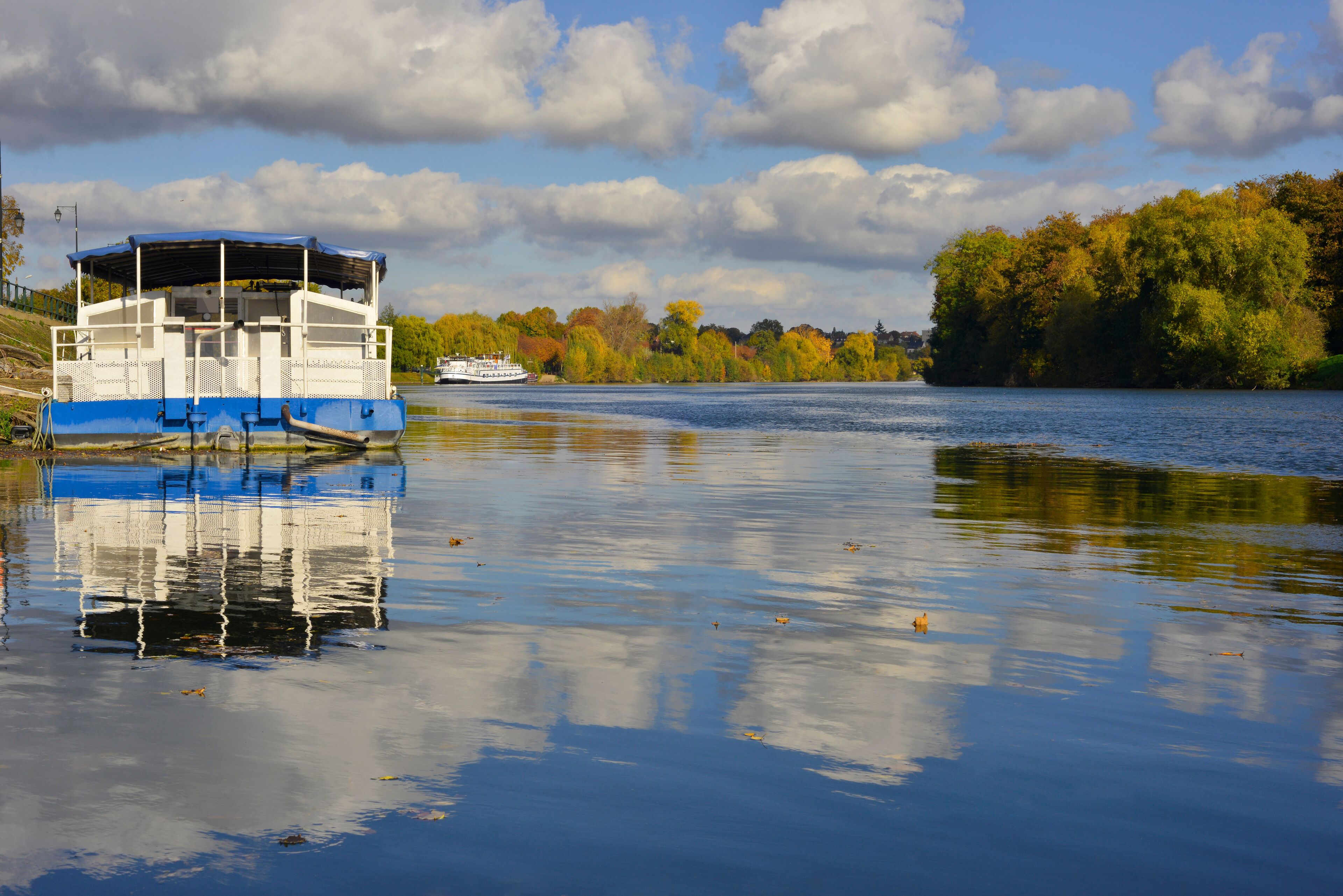 A river with a blue and white boat docked on the left and a white boat in the distance.