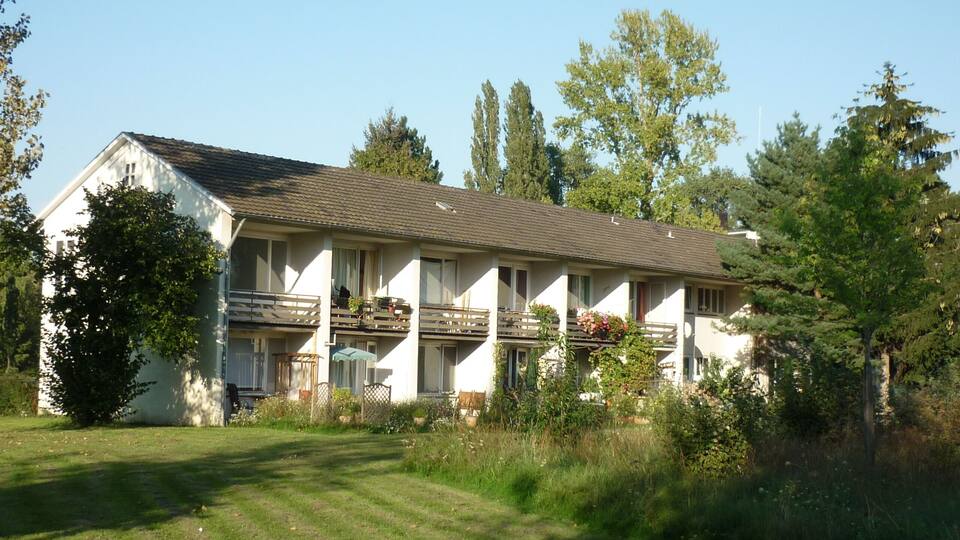 A two-story building with a sloped roof, surrounded by trees and a grassy area.