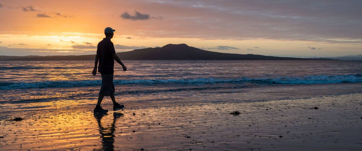 Silhouette man walking on Milford beach at sunrise, Rangitoto Island in the background, Auckland.