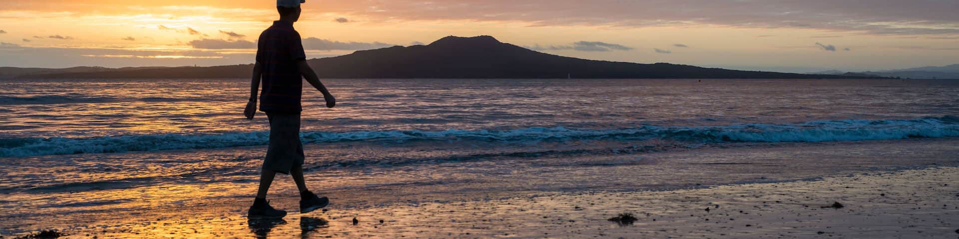Silhouette man walking on Milford beach at sunrise, Rangitoto Island in the background, Auckland.