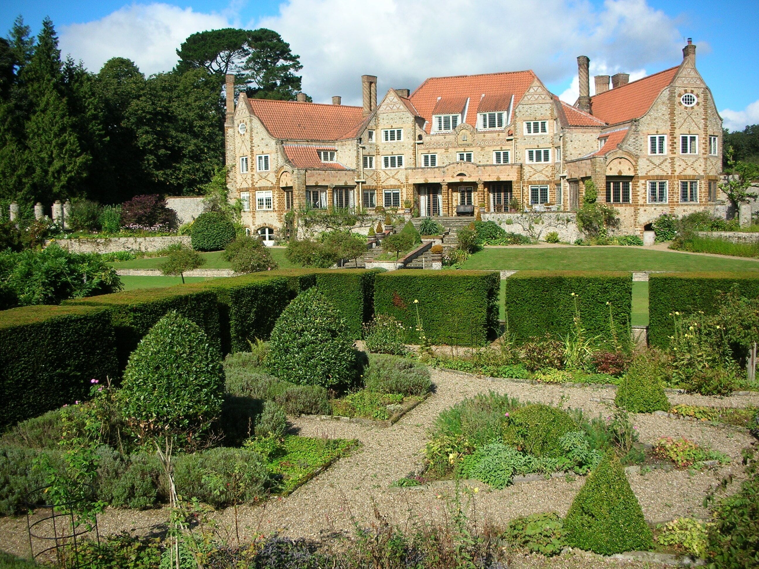 A large, historic stone building with a red-tiled roof, surrounded by well-manicured gardens and hedges.