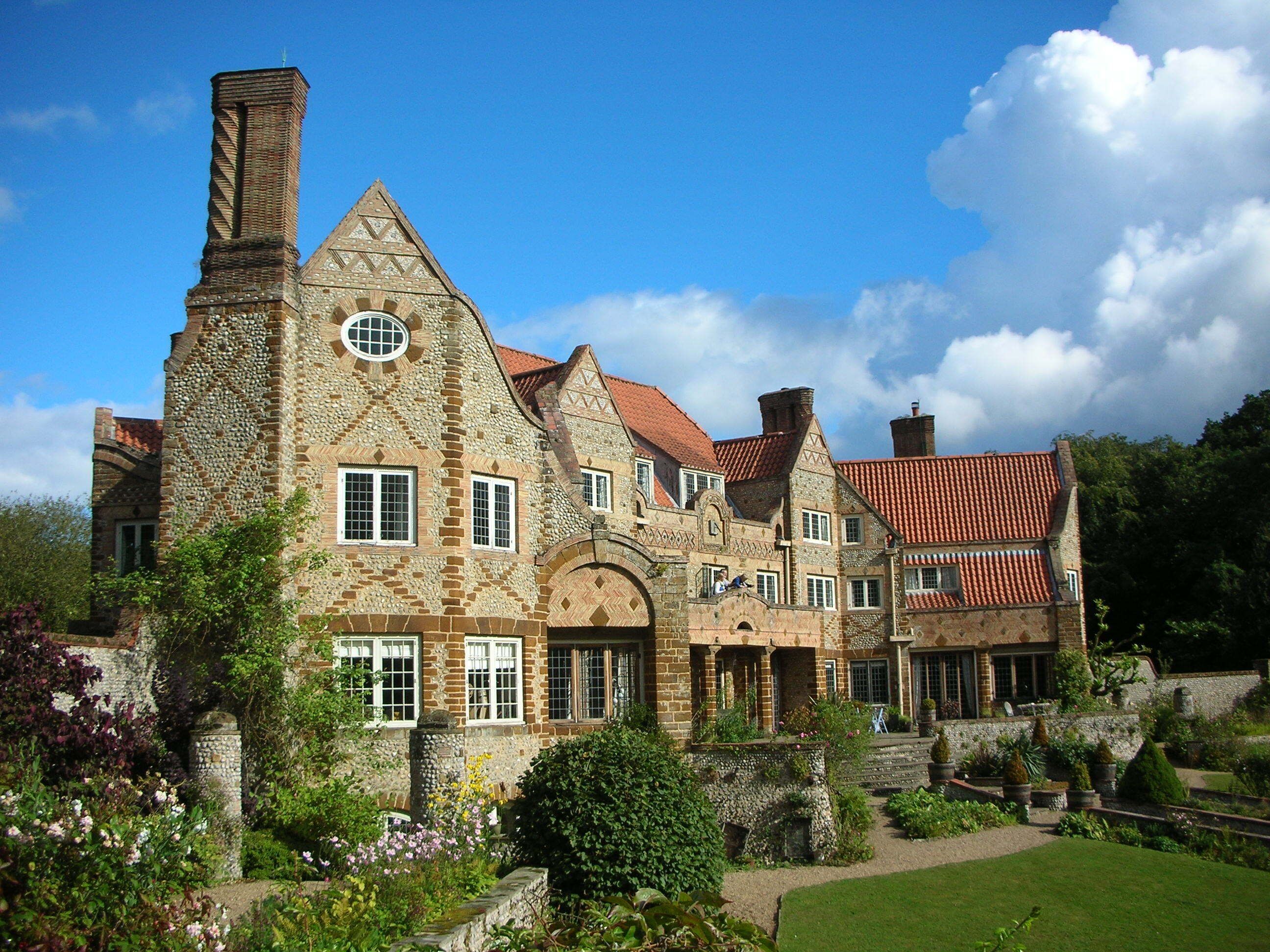 A large stone manor with a red-tiled roof, surrounded by lush gardens and a clear blue sky.