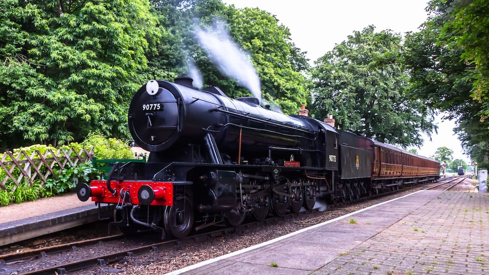 Une locomotive à vapeur portant le numéro 90775, sur une voie ferrée, avec une plateforme en briques et une végétation luxuriante en arrière-plan.