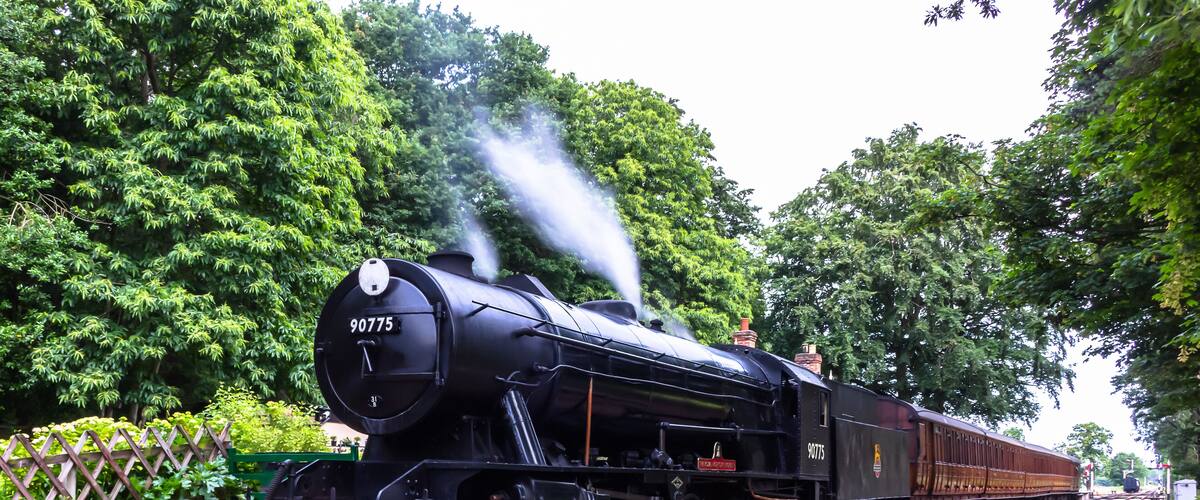 A steam locomotive numbered 90775 on a railway track with a brick platform and lush greenery in the background.
